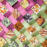Mixed Brownies assortment by Crumbs & Doilies featuring various flavors arranged on pink and green backgrounds.
